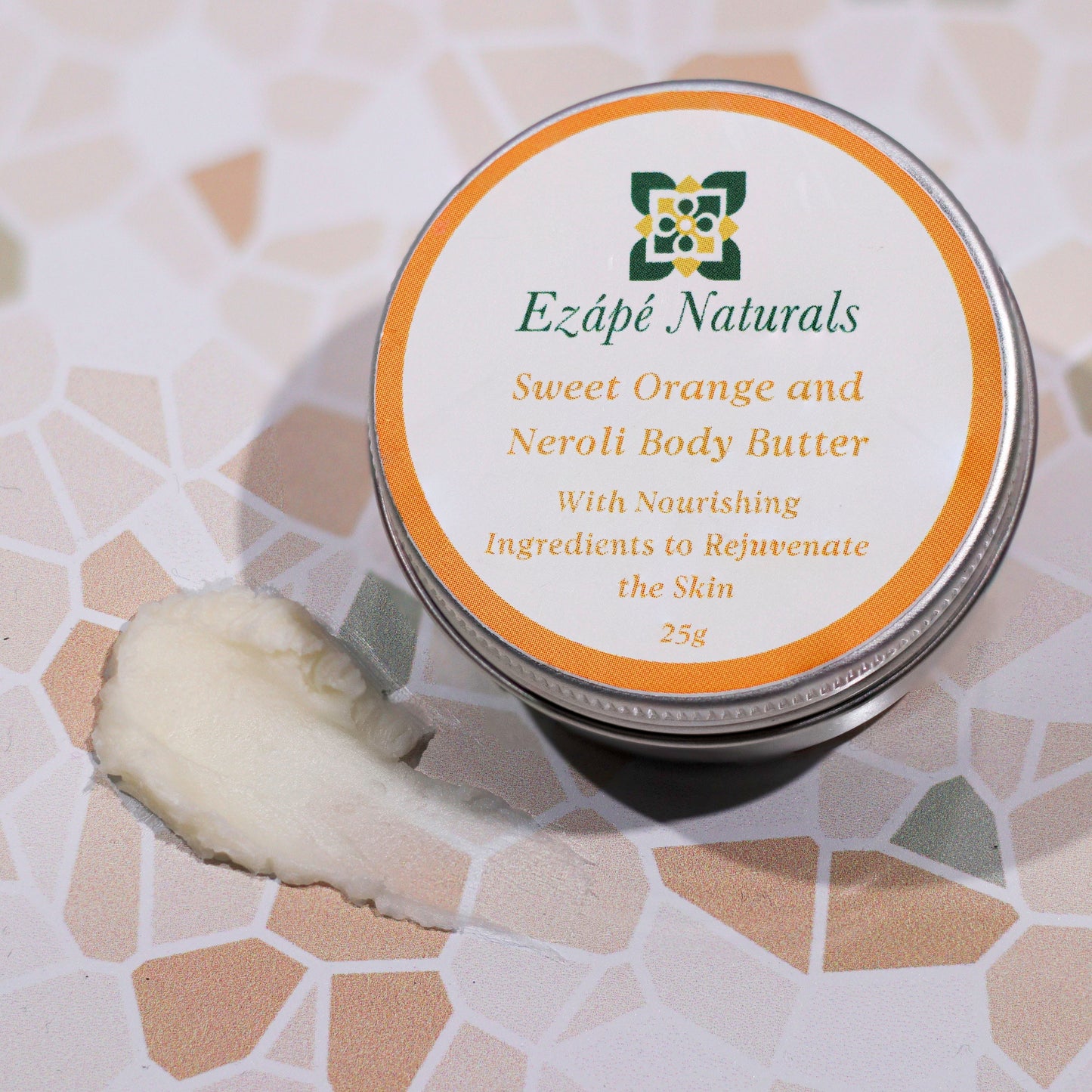 Ezape Naturals Sweet Orange and Neroli Body Butter on a textured surface that looks like a mosaic. there is a slick of body butter showing so you can see the texture and then then small round aluminium tin that it comes in which is labelled with a round white sticker with an orange border