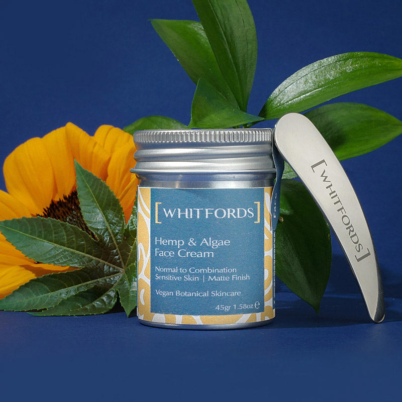 whitfords skincare hemp and algae face cream for normal to combination skin in an aluminium tin with a grey blue label on a royal blue background next to a stainless steel spatula, some waxy green leaves, a sunflower and a tropical plant green leaf.. the label reads 'whitfords hemp and algae face cream. normal to combination, sensitive skin, matte finish. vegan botanical skincare'