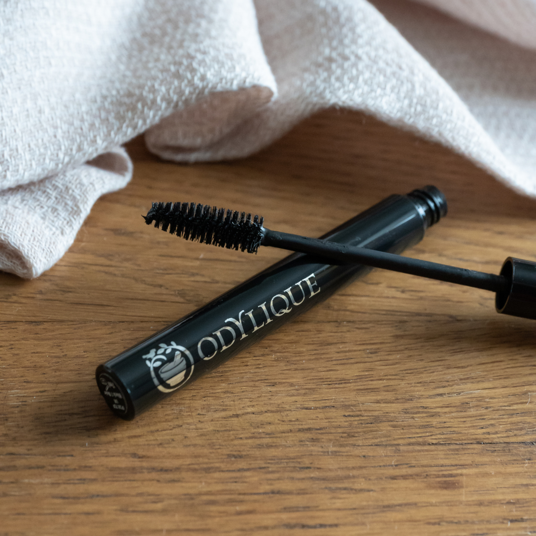 A tube of odylique mascara lying open on a wooden surface with its brush coated in mascara, near a softly folded white cloth.