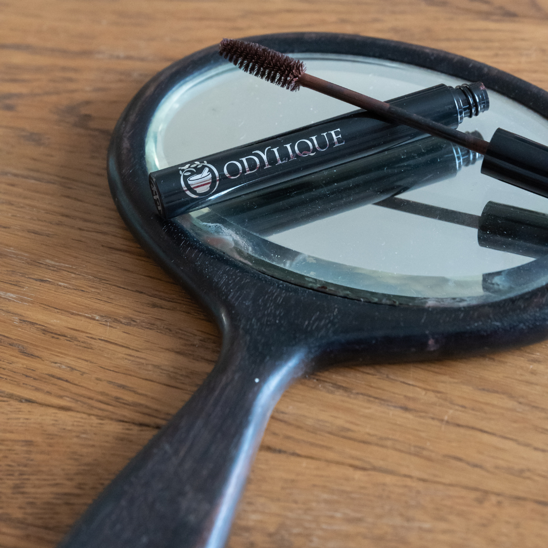 An old, oval hand mirror with a wooden handle lying on a wooden table, reflecting a mascara tube and brush from the brand 'odylique'.