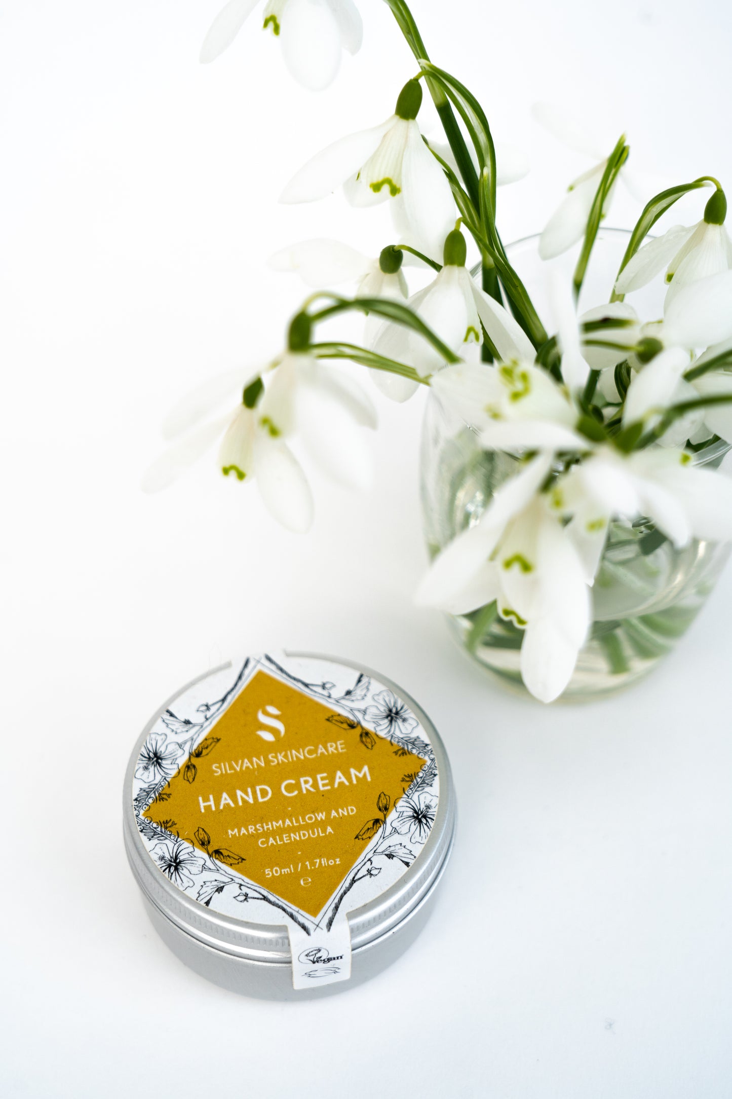 silvan skincare hand cream on a white background sitting next to a clear glass jar with water and some snowdrop flowers in the vase. the hand cream is in an aluminium pot with a white and yellow labels which reads: silvan skincare hand cream marshmallow and calendula 50ml