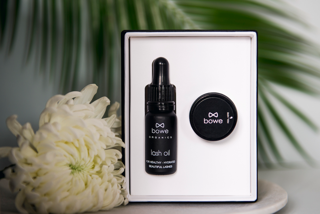 Bowe Organics Gift of Lips and Lashes. Natural Valentines gifts. The lash oil is pictured next to the lip balm in its box, with white flower next to it and a palm frond out of focus in the background.