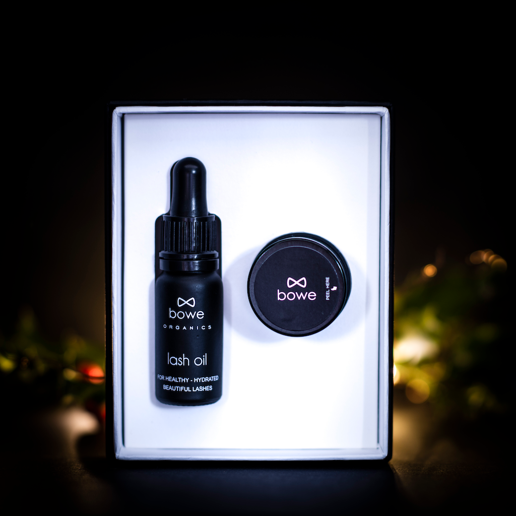 Bowe Organics Gift of Lips and Lashes. Vegan Christmas gifts. The lash oil and lip balm are pictures in their box, infront of a dimly lit Christmas background.