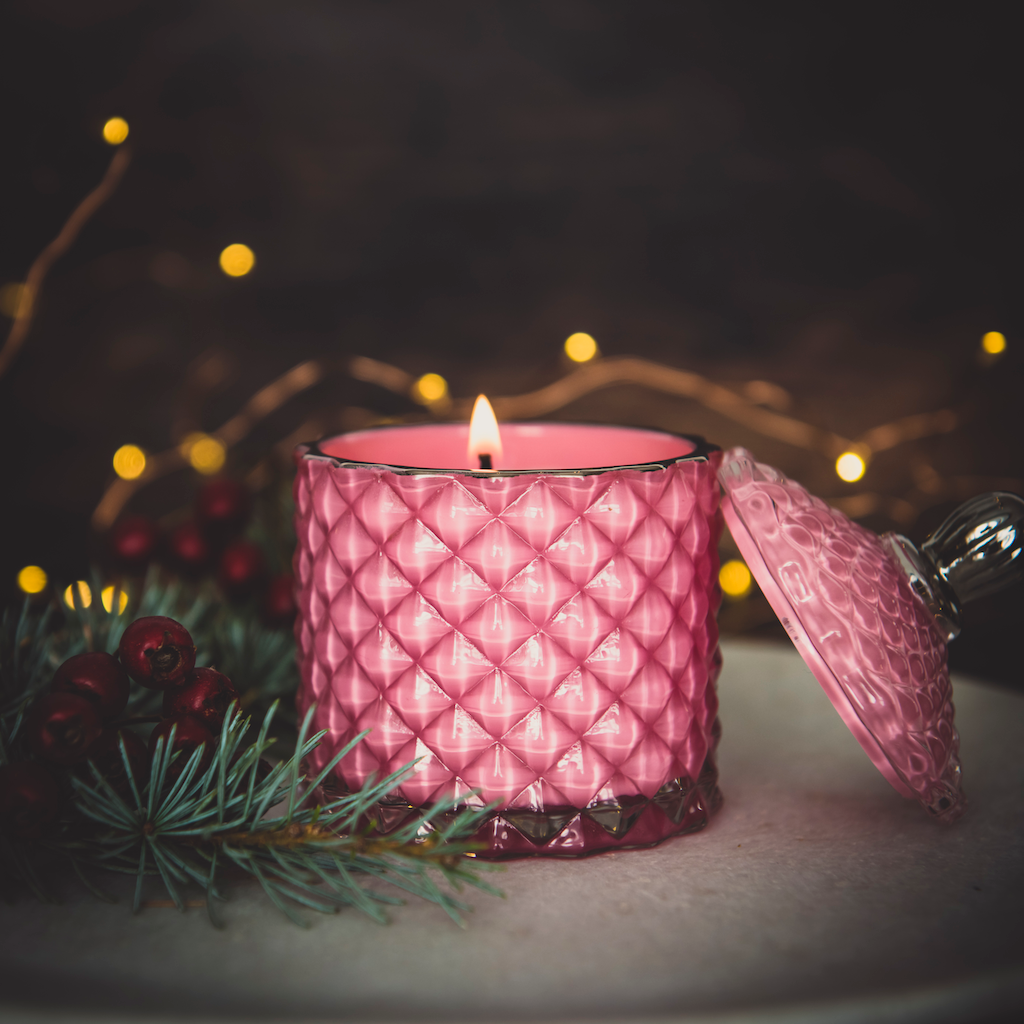 pink glass candle jar with lid off aflame with pine branch and twinkling lights