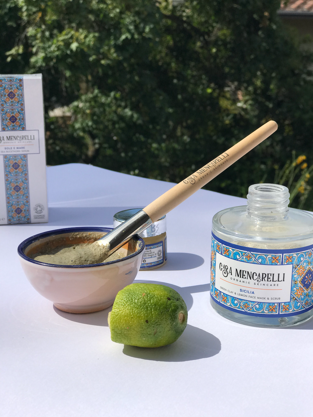 Casa Mencarelli Sicilia Green Clay & Lemon Face Mask & Scrub. Vegan face exfoliator. The glass jar is pictured open next to a mask bowl with a mask brush and the powder inside the bowl. A squeezed lime is sitting in front of the bowl.