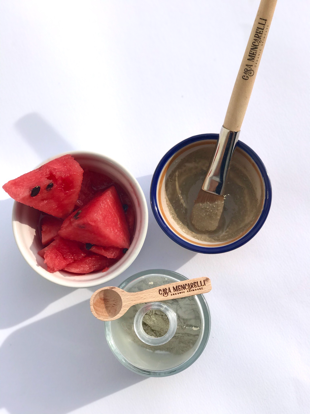 Casa Mencarelli Sicilia Green Clay & Lemon Face Mask & Scrub. Vegan face mask. The jar of mask is pictured open from above, next to a mask bowl with a brush and some of the mask in it, and another bowl with small cubes of watermelon.