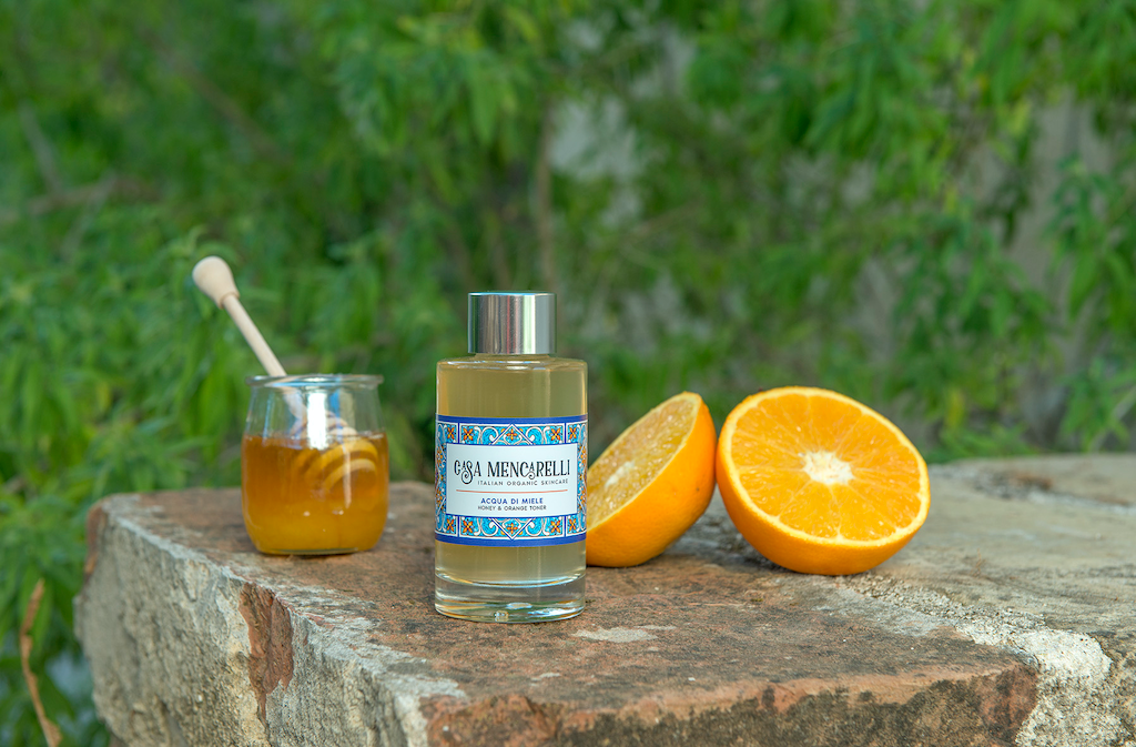 Casa Mencarelli Acqua di Miele Honey & Orange Toner. Organic toner. The toner bottle is pictured on a stone wall with a sliced orange next to it and a little jar of honey.