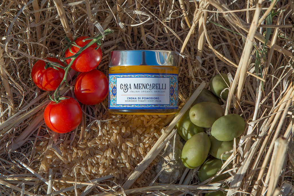 Casa Mencarelli Crema Di Pomodoro Nourishing Tomato Night Cream & Cleanser. Natural, multi-purpose skincare products. The product is packaged in a glass jar with a label decorated with images of the traditional tiling of Umbria. It is sitting in a bunch of hay with whole grain rice, tomatoes and grapes next to it.