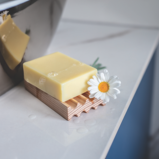 creamy coloured chamomile soap bar resting on a wooden soap tray on the side of a sink. You can see the reflection of the soap in the basin