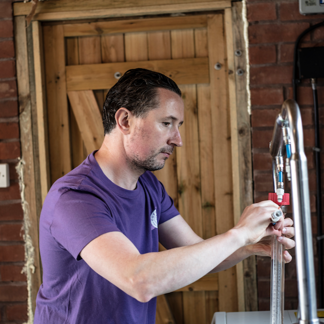 kevin, coraline skincare's founder working in his soapery in devon. he is distilling some essential oils and behind him is a red brick wall with wooden panelled door