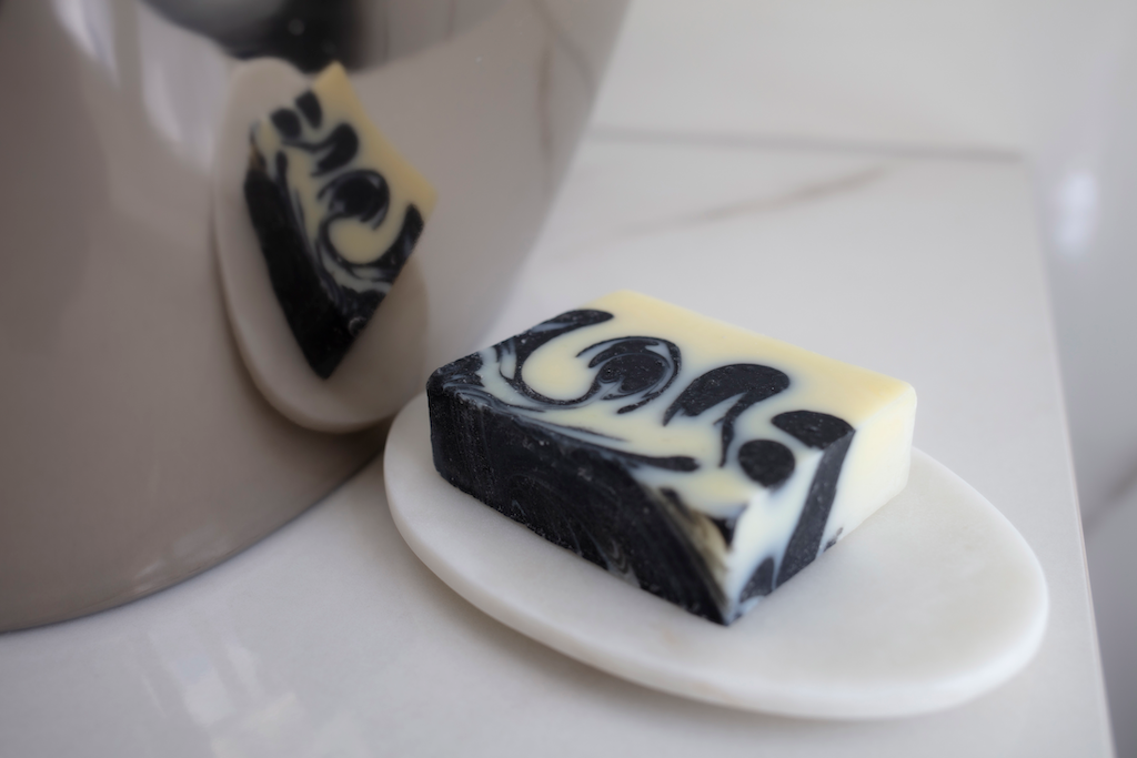 A bar of the minty fresh soap rests on a white soap dish, which sits on a smooth, light-colored surface. The soap's reflection can be seen in a shiny, curved surface nearby.