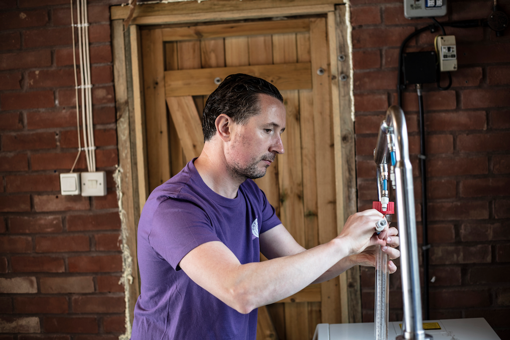 kevin, coraline skincare's founder working in his soapery in devon. he is distilling some essential oils and behind him is a red brick wall with wooden panelled door