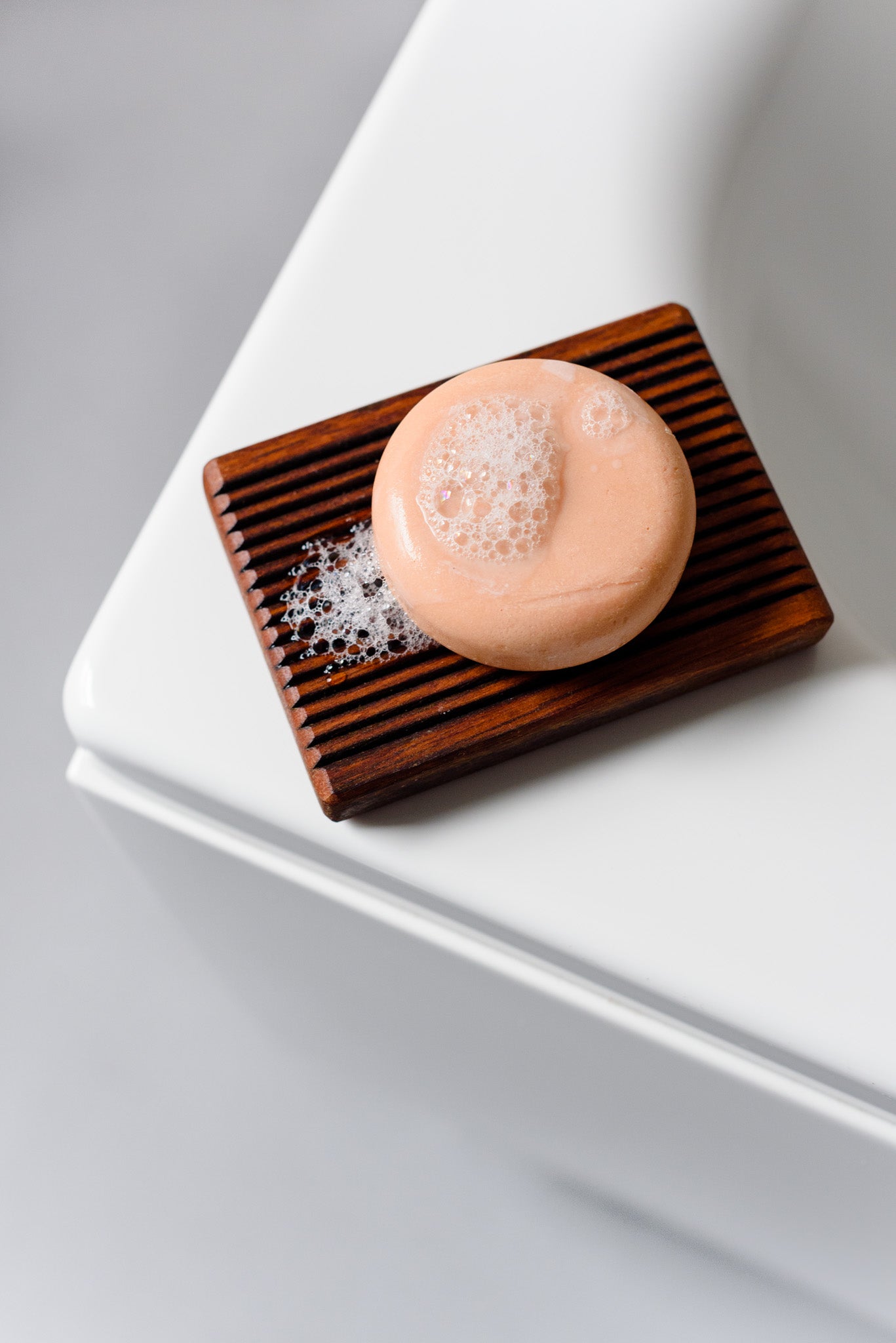 The round pink shampoo bar, foamy and bubbly, placed on a wooden soap tray, resting on a white shelf.