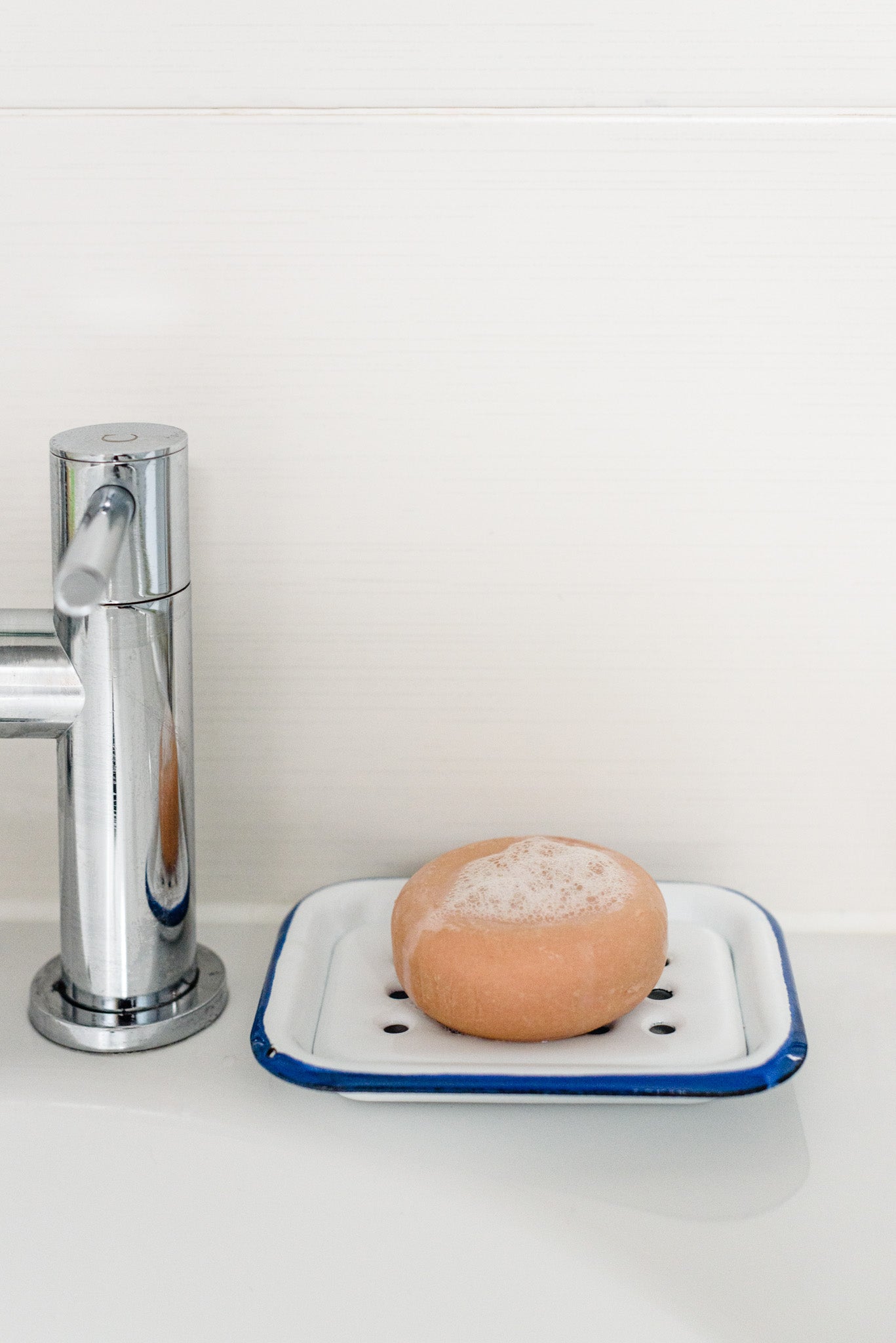 The round pink shampoo bar, foamy and bubbly, placed on an enamel tray, resting on a white sink next to a silver tap.