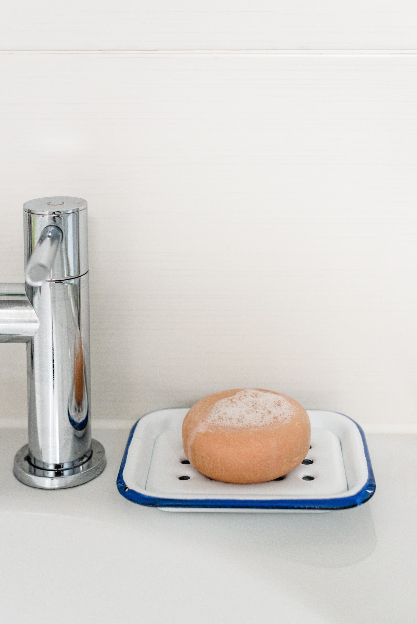 The round pink shampoo bar, foamy and bubbly, placed on an enamel tray, resting on a white sink next to a silver tap.
