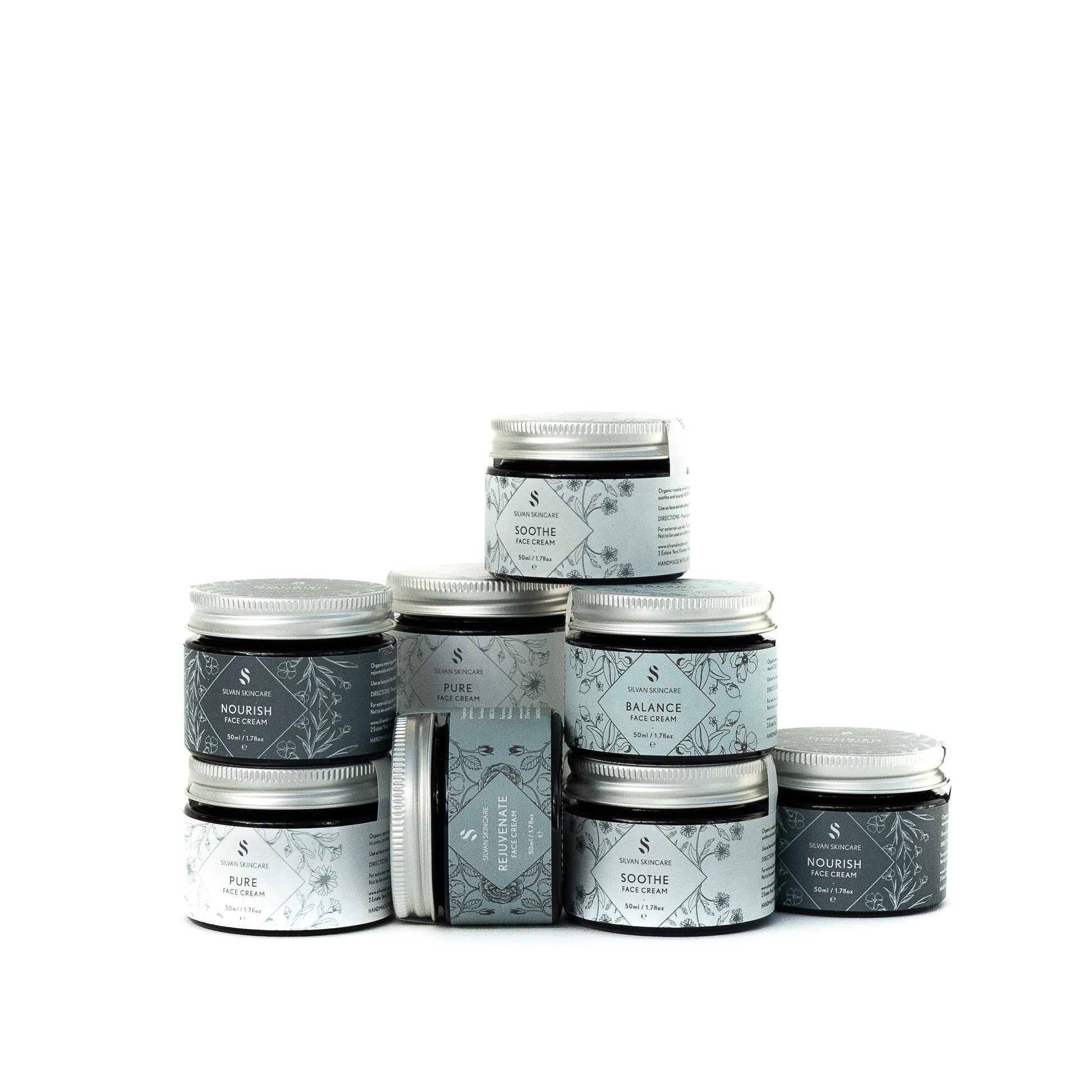 Several jars of the Silvan faces creams are arranged in a pyramid like formation. The jars are labelled in different shades of blue, indicating the different creams. Each jar has a silver aluminium lid and botanical illustrations.