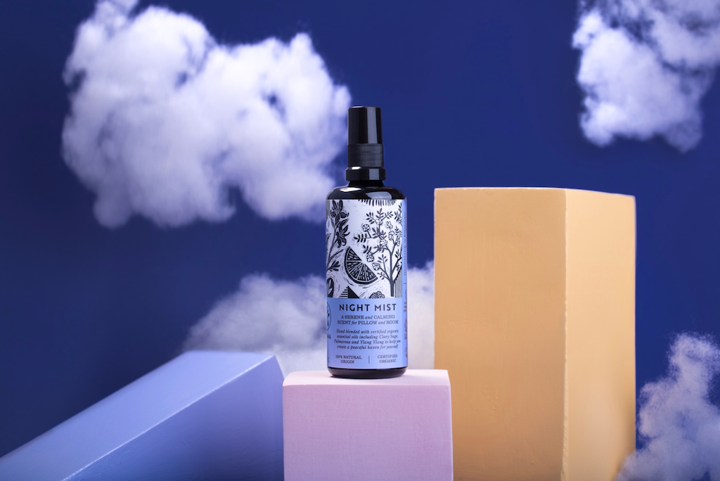 A night mist spray bottle is positioned among pastel-colored rectangular blocks against a deep blue background. The backdrop features several fluffy white clouds, creating a dreamy, whimsical setting.