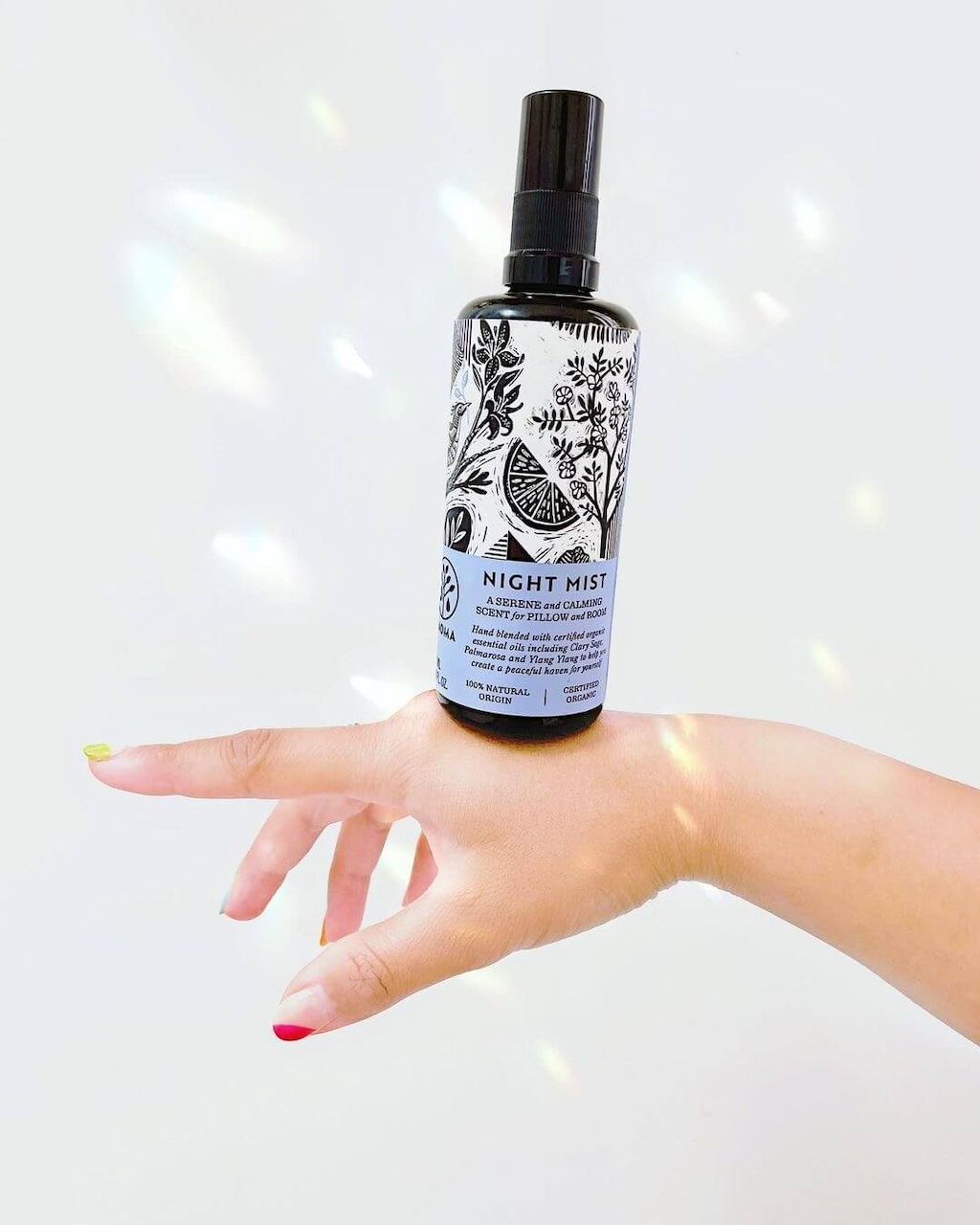 A hand with multicolored nails holds a bottle of "Night Mist" facial toner. The bottle has a black spray cap and a label featuring botanical illustrations. The background is plain white with light reflecting off the walls providing a minimalistic and clean appearance.