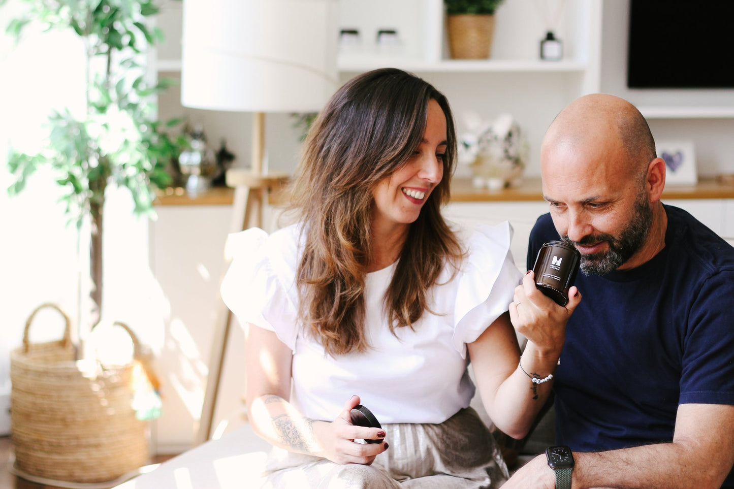 julieta and adrien, founders of holistic london are sitting in a bright cost room with white shelving in the background and a plant in a wicker basket. julieta is encouraging adrien to smell one of their natural vegan candles in a glass jar