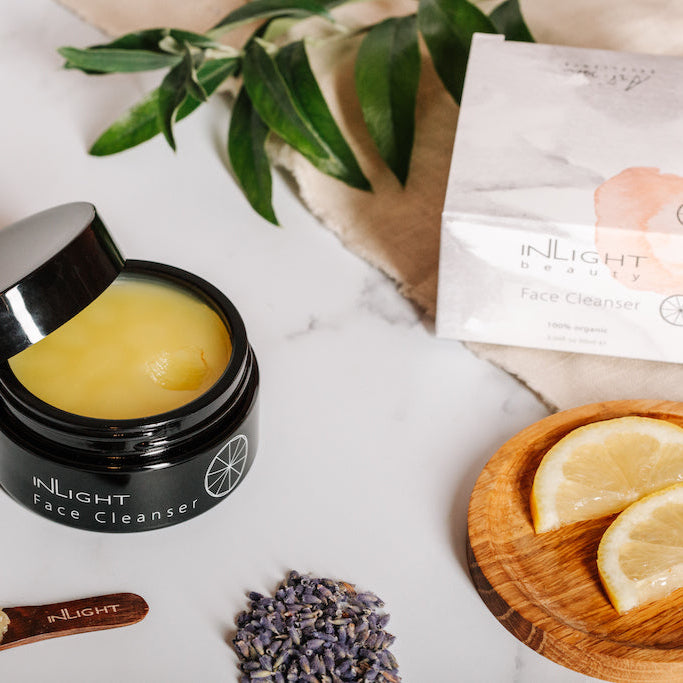 flatlay of inlight beauty's face cleanser balm. the open pot of cleanser can be seen surrounded by some fresh ingredients that are in the product. 2 slices of lemon on a wooden coaster, some dried lavender in a pile and also on the stem and a sprig of bay leaves. there is an organic fabric laying under the leaves which the box for the cleanser is sitting on