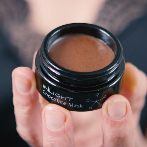 image looking down in to an open pot of the inlight beauty 100% organic chocolate mask that's hand made in cornwall. The jar is black and is being held in someone's finger tips. The mask is a rich warm brown colour. In the background is a blurred image of the persons black top and decollotege