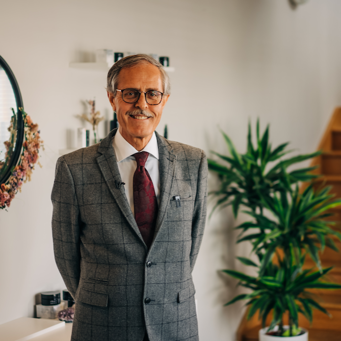 dr spiezia founder of inlight beauty who is standing in a grey checked suit in a hallway with white paint, a black mirror surrounded by pink flowers and a spiky green palm plant and pine coloured stairs in the background