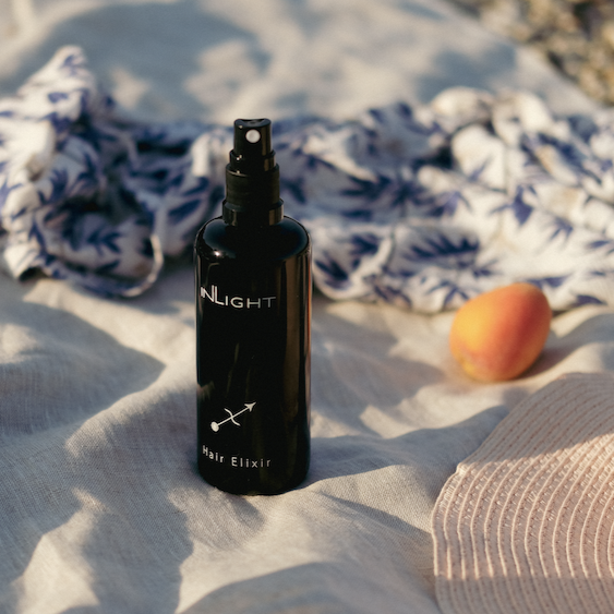 inlight beauty organic hair oil lifestyle photo showing the black miron glass bottle of hair elixir sitting on a gauzy linen fabric in dappled sunshine next to the textured fabric of a wicker hat, an apricot and a scrumpled up scarf or shawl with a blue palm print blurred in the background