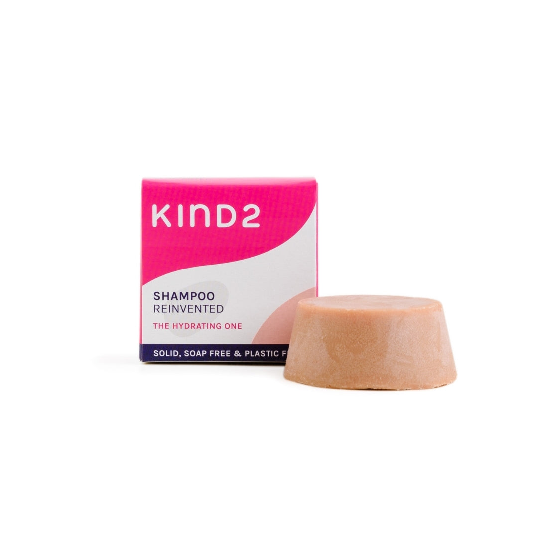 A small round pink plastic free KIND2 shampoo bar. Placed in front of its small pink, white and navy box. 