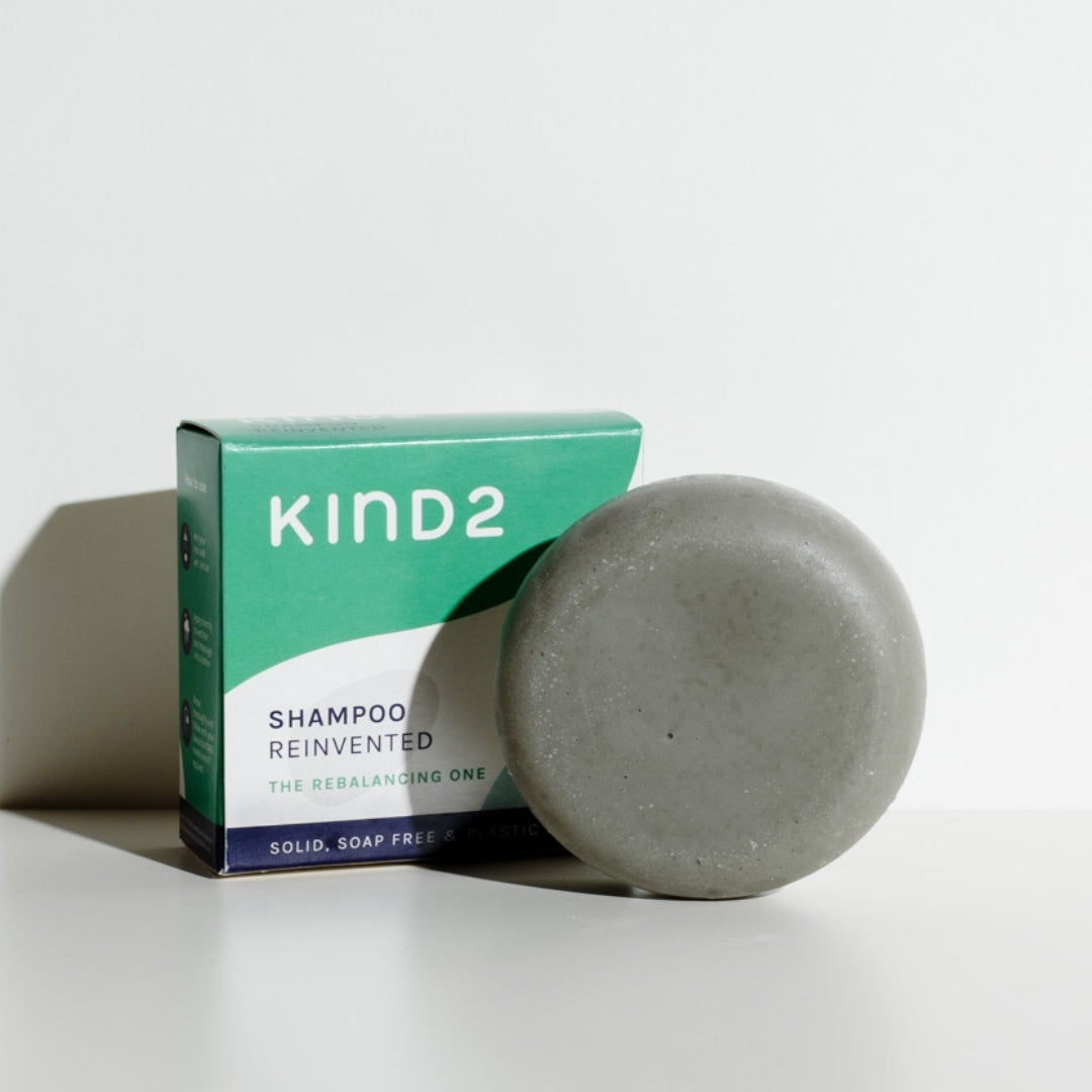 A circular grey KIND2 shampoo bar. Placed next to the green and white it comes in.