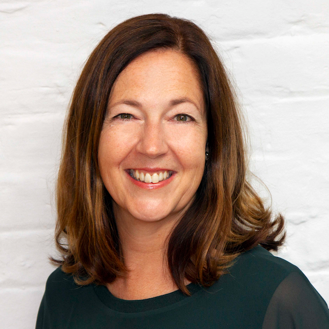 A picture of KIND2 founder Sue Campbell smiling. She is dressed in dark green and stood in front of a white brick wall.