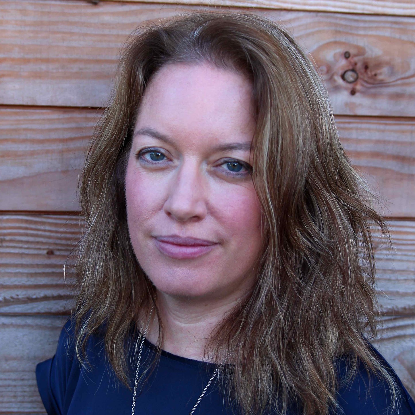 photo of marian who makes silvan skincare vegan face creams from her studio in the welsh borders. Photo of marian standing in front of natural wood panelling with light brown hair and a blue boat neck top with a gold chain