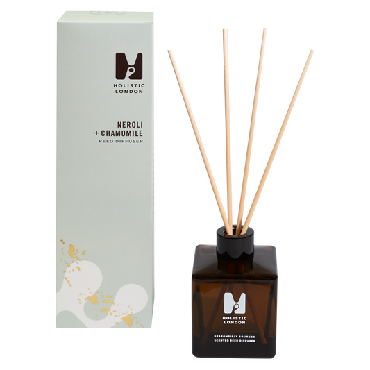 photograph of holistic london's neroli + chamomile reed diffuser on a white background. on the left side of the image is the light green cardboard box with the holistic london logo on it. the label reads ' neroli + chamomile reed diffuser’. on the right side of the photo is the amber glass bottle with 4 wooden reeds inside. the glass is printed with the holistic london logo and says 'responsibly sourced scented reed diffuser’