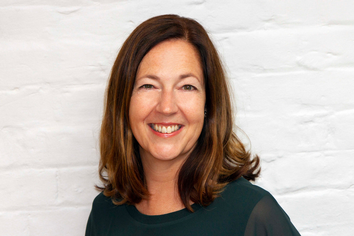 A picture of KIND2 founder Sue Campbell smiling. She is dressed in dark green and stood in front of a white brick wall.