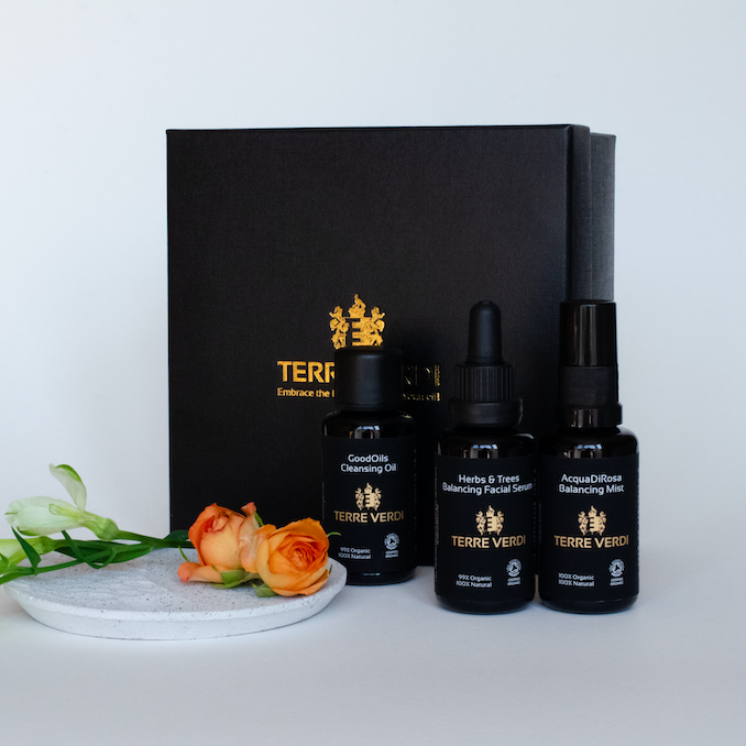 Terre Verdi Organic Gift Set for Face. Natural skincare gift set. All of the products are in black bottles and stand next to a luxury black box embossed in gold with the Terre Verdi logo. There are green flower buds and orange roses on a plate in front of the box.