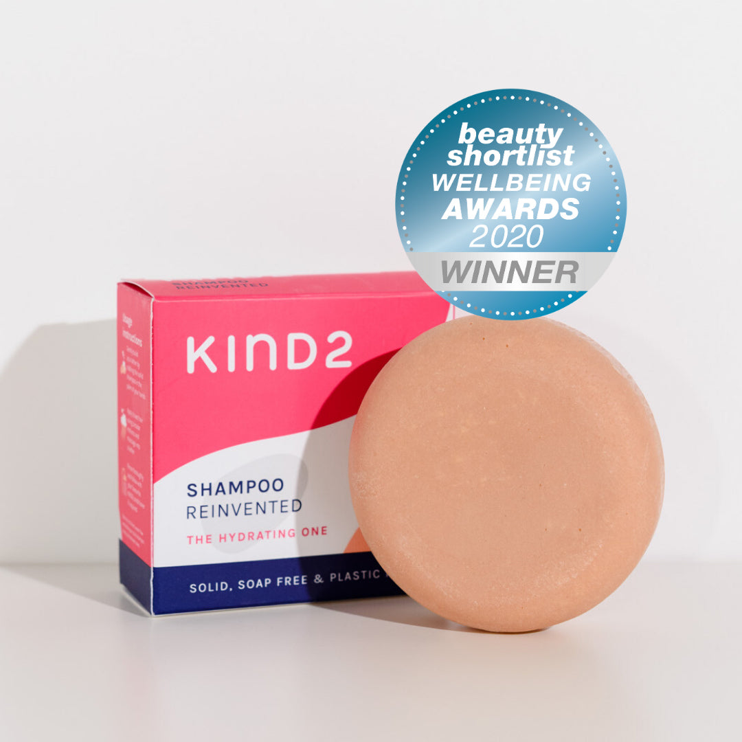 Round pink plastic free KIND2 shampoo bar. Placed in front of its pink, white and navy box. A Beauty Shortlist Wellbeing Awards 2020 Winner Sticker on top of them.