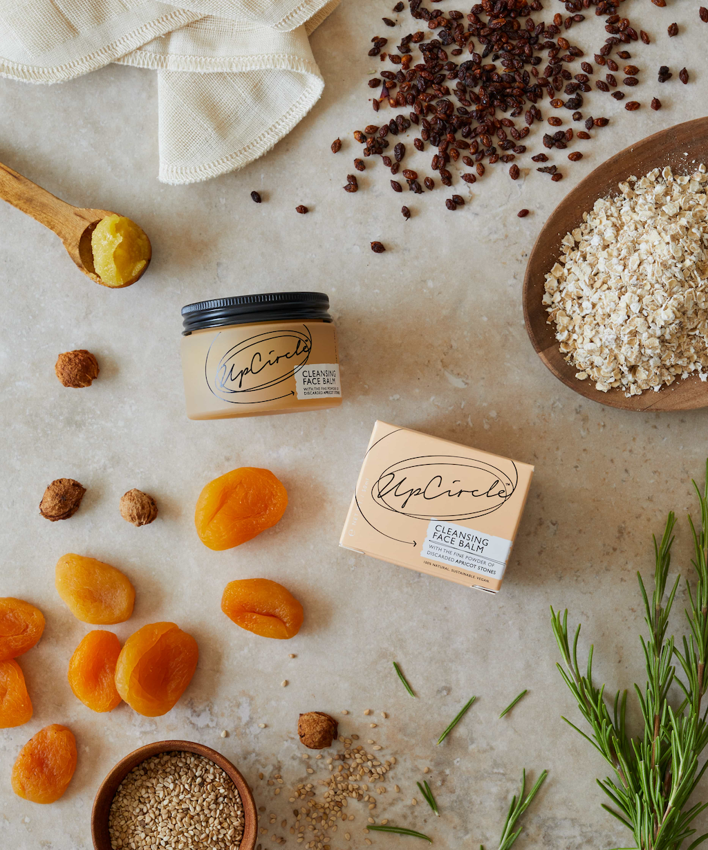 UpCircle Cleansing Face Balm. Sustainable skincare. The orange glass jar containing the balm is sitting next to the branded orange box it comes in. They are surrounded by the various ingredients that make up the cleansing balm.