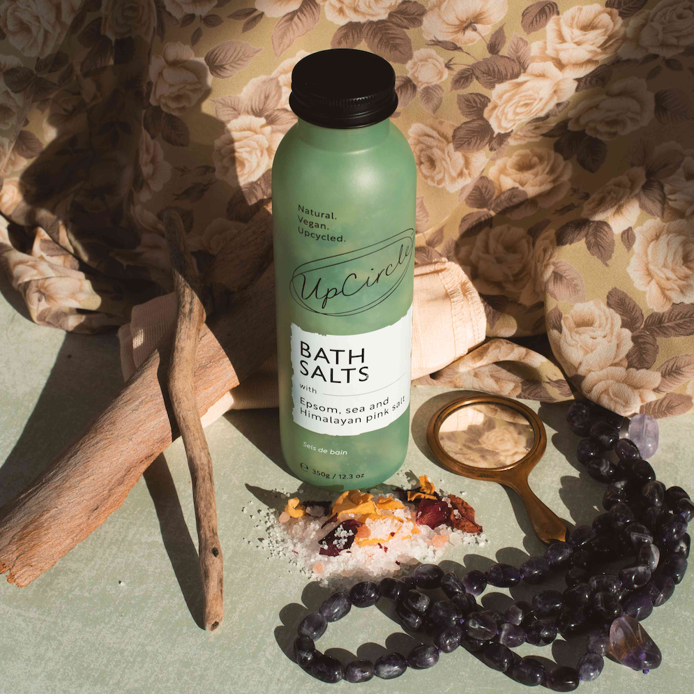 upcircle bath salts being displayed in a vintage style setting with floral brown tonal fabric behind and a small bronze hand mirror and amethyst beads in the foreground next to some loose bath salts