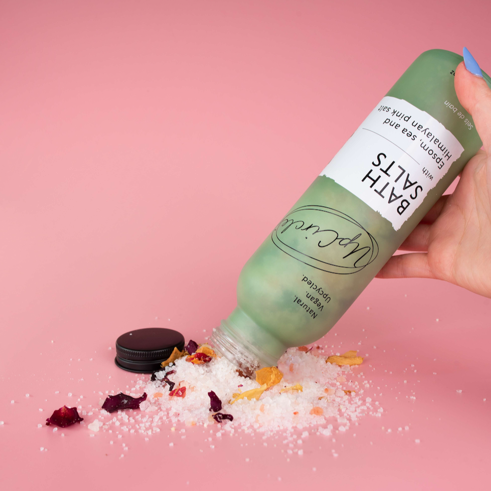 white and pink coloured bath salts and dried red and orange flower petals being poured out of a light sage green glass bottle onto a pink background