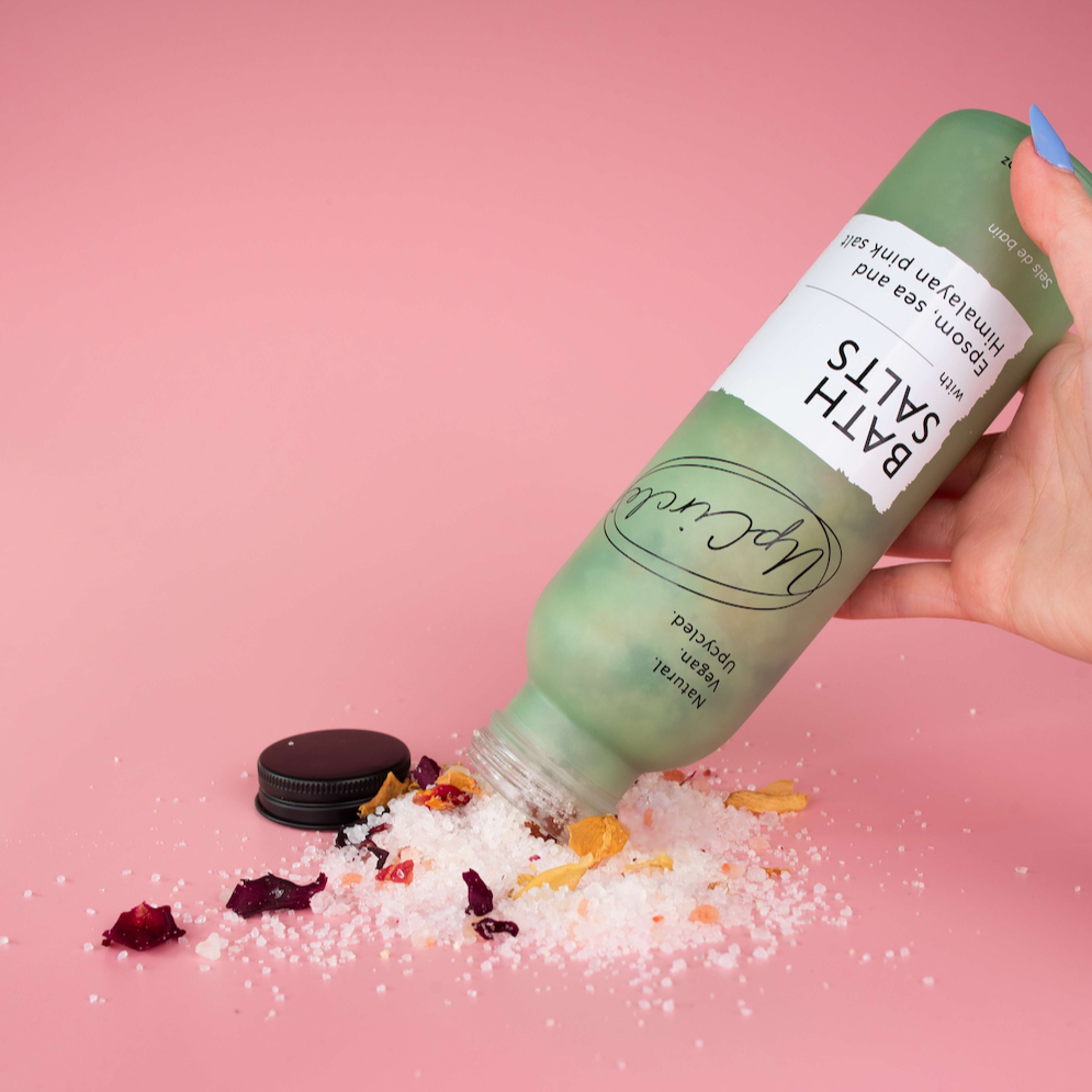 white and pink coloured bath salts and dried red and orange flower petals being poured out of a light sage green glass bottle onto a pink background 