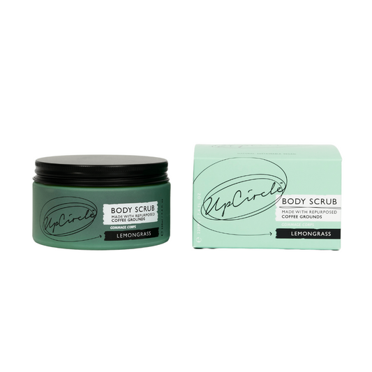 A green glass jar of UpCircle Lemongrass Body Scrub sits on a white background. It has a black aluminium lid and is placed next to the plastic free packaging box it comes in, on the right.