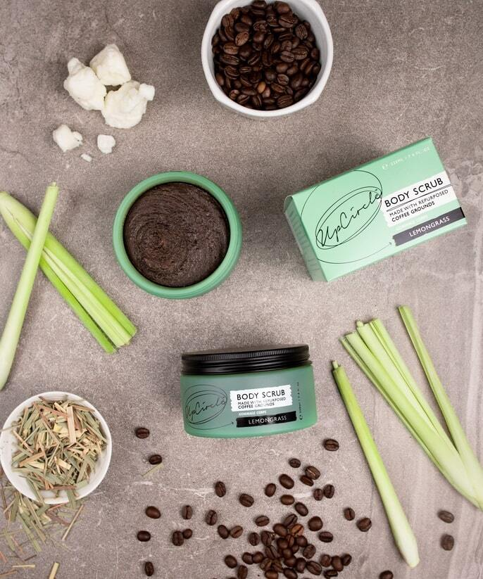An arrangement of the products to make the upcircle body scrub are displayed on a grey surface. Items include the jar of the scrub, a bowl of coffee beans, cut lemongrass stalks, and a bowl with lemongrass pieces. Coffee beans and lemongrass pieces are scattered around.