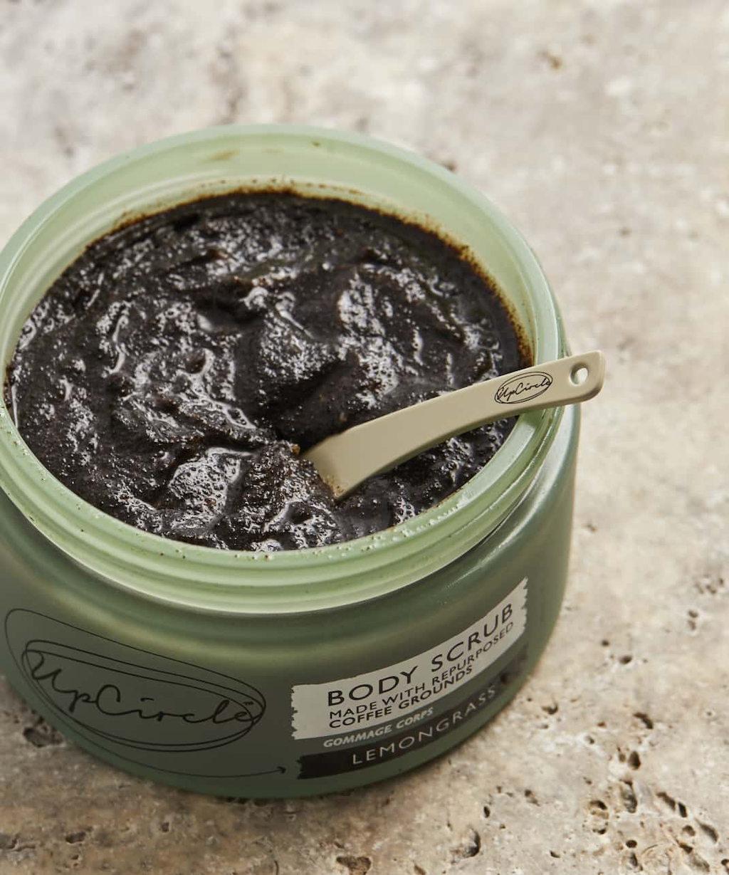 The upcircle body scrub is placed on a stone surface. The image has been taken from above with the jar open, with an upcircle spatula placed inside.