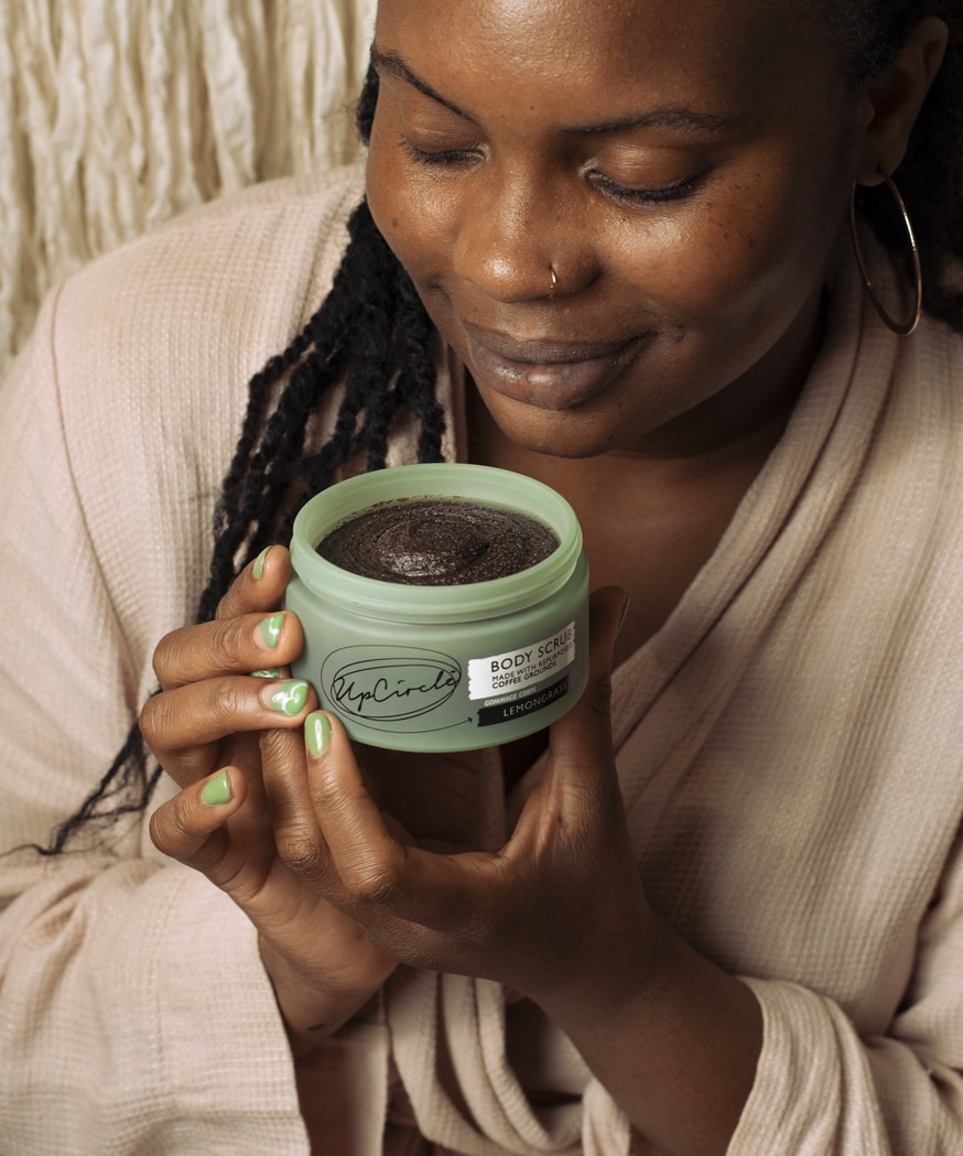 An image of a woman holding the body scrub in her hands. She is dressed in a beige robe and has her eyes closed as she takes in the scent of the scrub.
