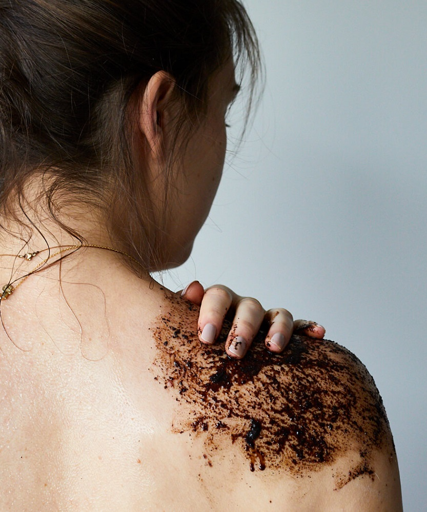 An image of a woman with her hair loosely tied up. She has her back to camera and is shown using the scrub on her right shoulder and massaging it in with her hand.