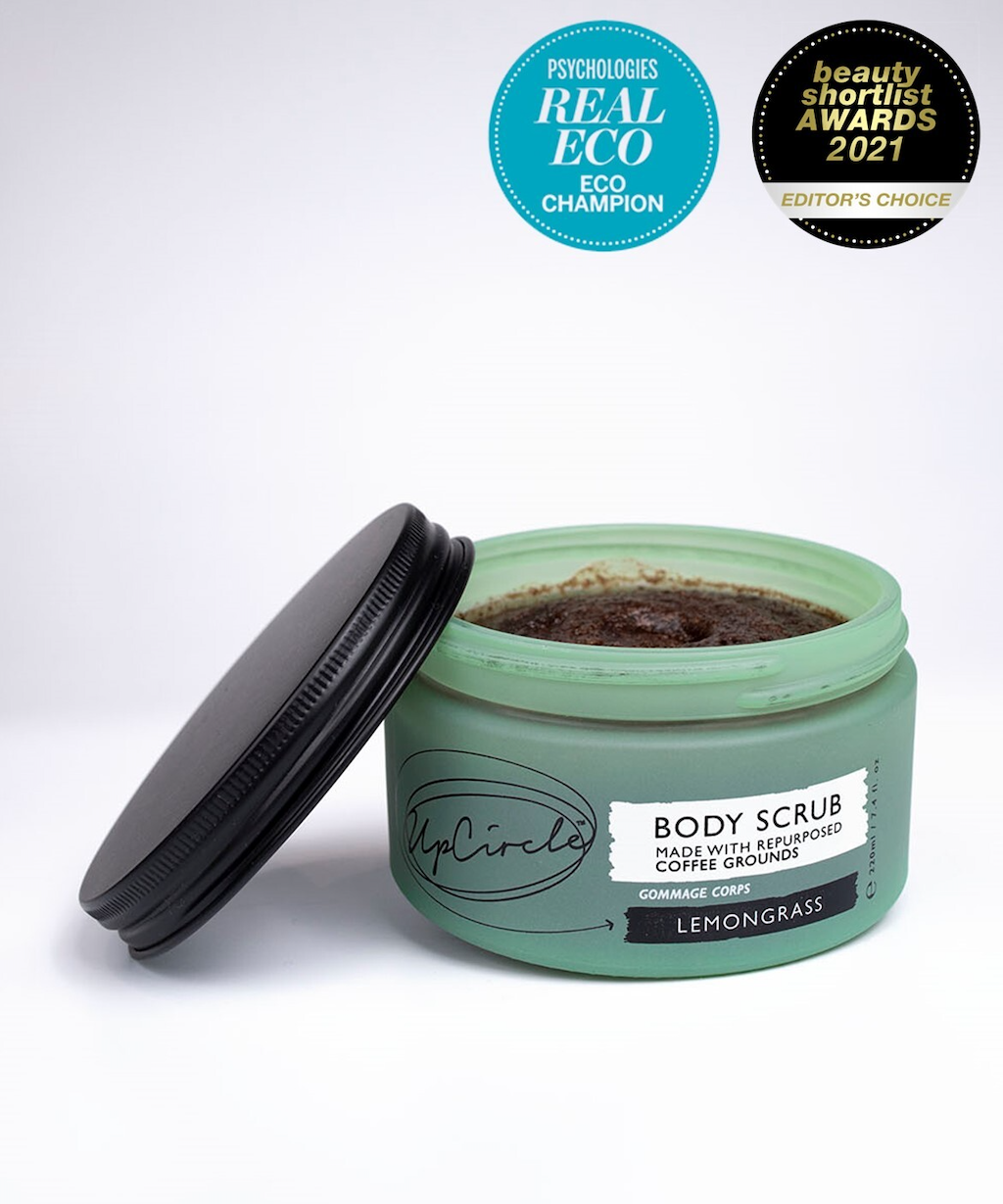 A green glass jar of UpCircle Lemongrass Body Scrub made with repurposed coffee grounds sits on a white background. The lid is off, revealing the scrub inside. Award badges for "Psychologies Real Eco Champion" and "Beauty Shortlist Awards 2021 Editor's Choice" are displayed above.