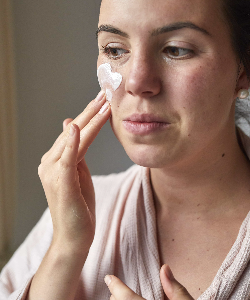 white woman with brown hair in a light blush pink robe applying upcircle face moisturiser