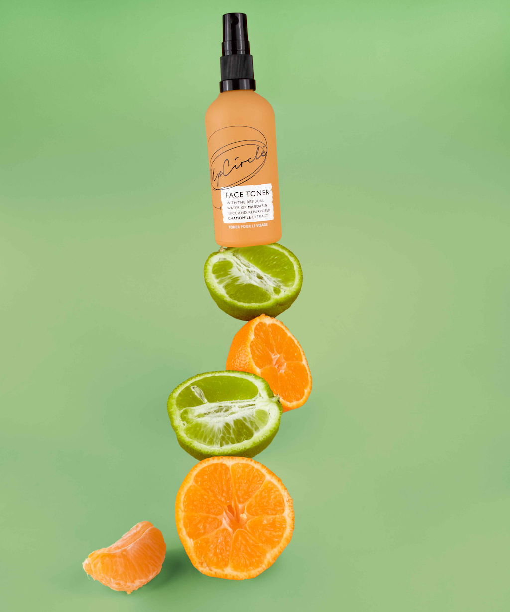 upcircle face toner bottle on a minty green background is balancing on a stack of orange and lime segments