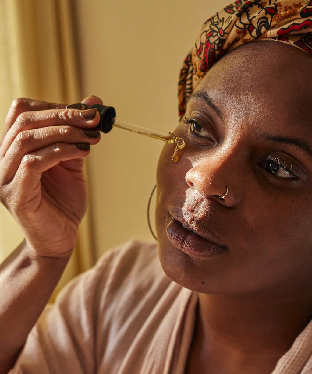 An image of a woman using the face oil She is holding the pipette in one hand as she places the product on to her skin. We can see a drop of serum as it spills from the pipette and down her face.