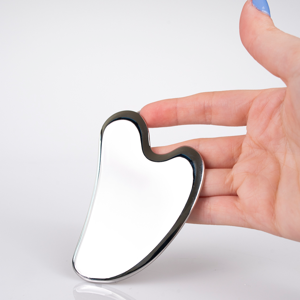 A hand with light skin and blue-painted nails holds a shiny, metallic Gua Sha tool with smooth, curved edges against a plain white background.