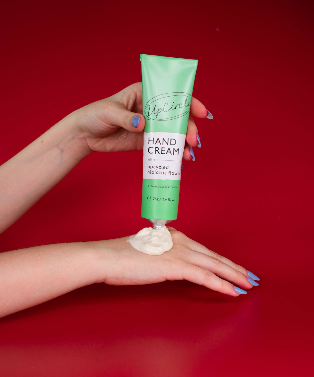 hands with blue nail polish is squeezing a green tube of UpCircle hand cream with upcycled hibiscus flower onto the back of their hand. The background is a rich red color, making the product and hands stand out.